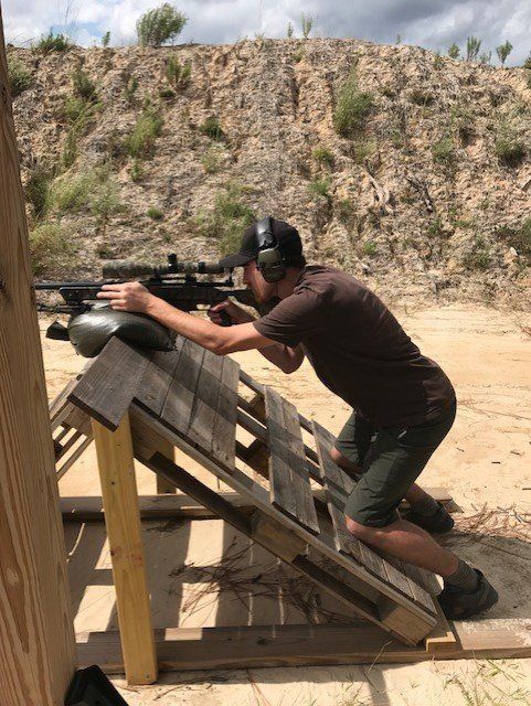 Man kneeling at a shooting range, aiming a rifle resting on a wooden support.