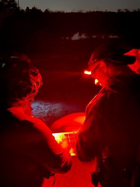 Two people at night, illuminated by red lights. One looks at a screen, the other stands nearby.