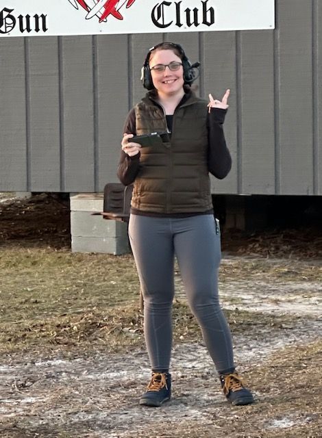 Woman at a gun range wearing ear protection and a vest, holding a tablet, giving a rock-on hand gesture.