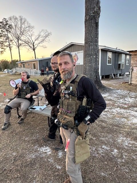Three men in tactical gear outdoors; one waves with a smirk, two others in background.