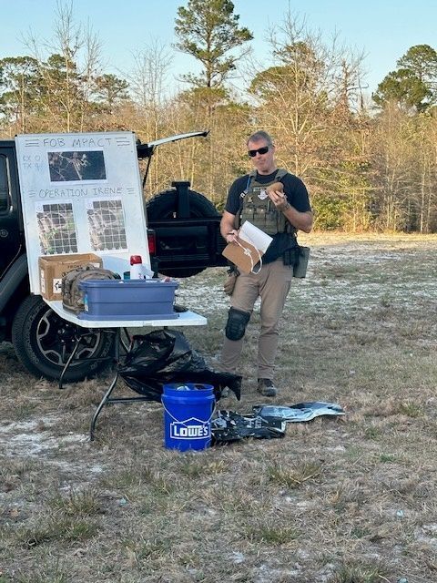A law enforcement officer stands by a table with evidence, a sign. Outdoors, sunny.