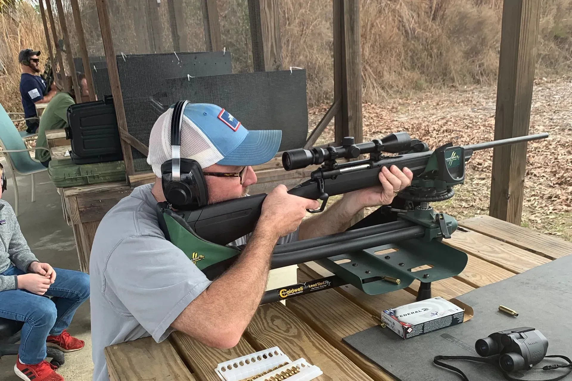 Man aiming a rifle at an outdoor shooting range, wearing ear protection, a blue hat, and glasses.
