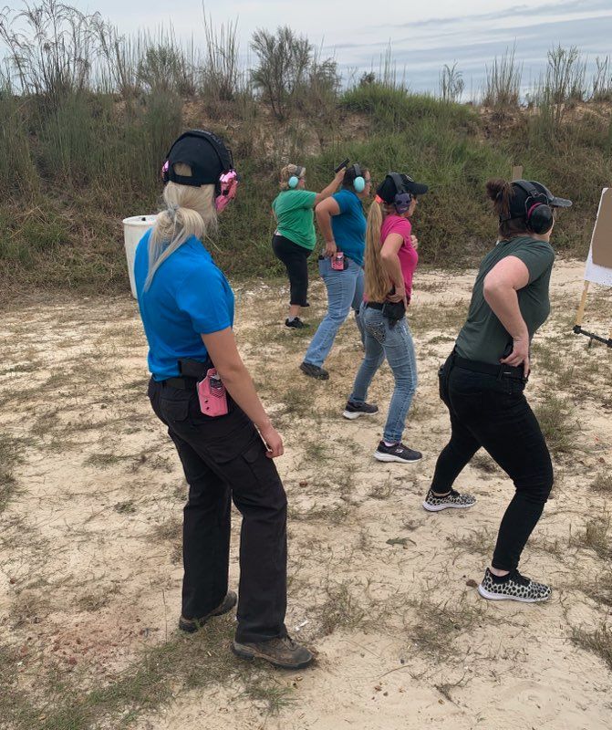 Women at an outdoor shooting range, practicing pistol shooting. They wear ear protection and hold guns.