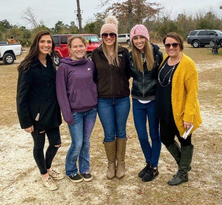 Five women pose outside near a red Jeep. They wear winter clothing in a field with light-colored sand.