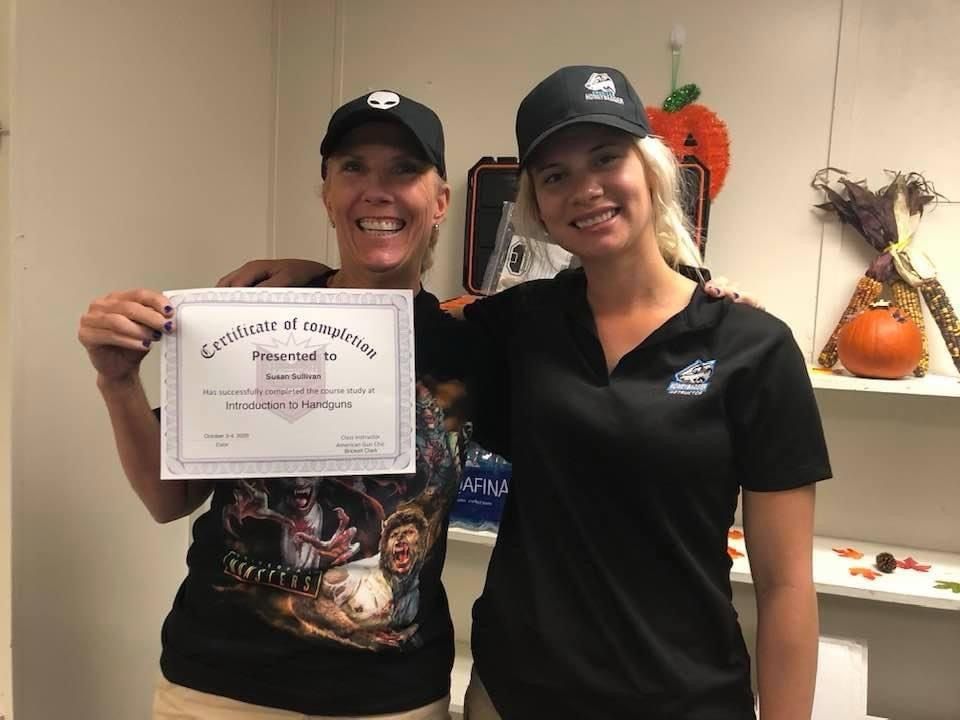 Two women holding a certificate. One wears a black cap and shirt, the other a black cap and graphic tee. A festive setting is visible.