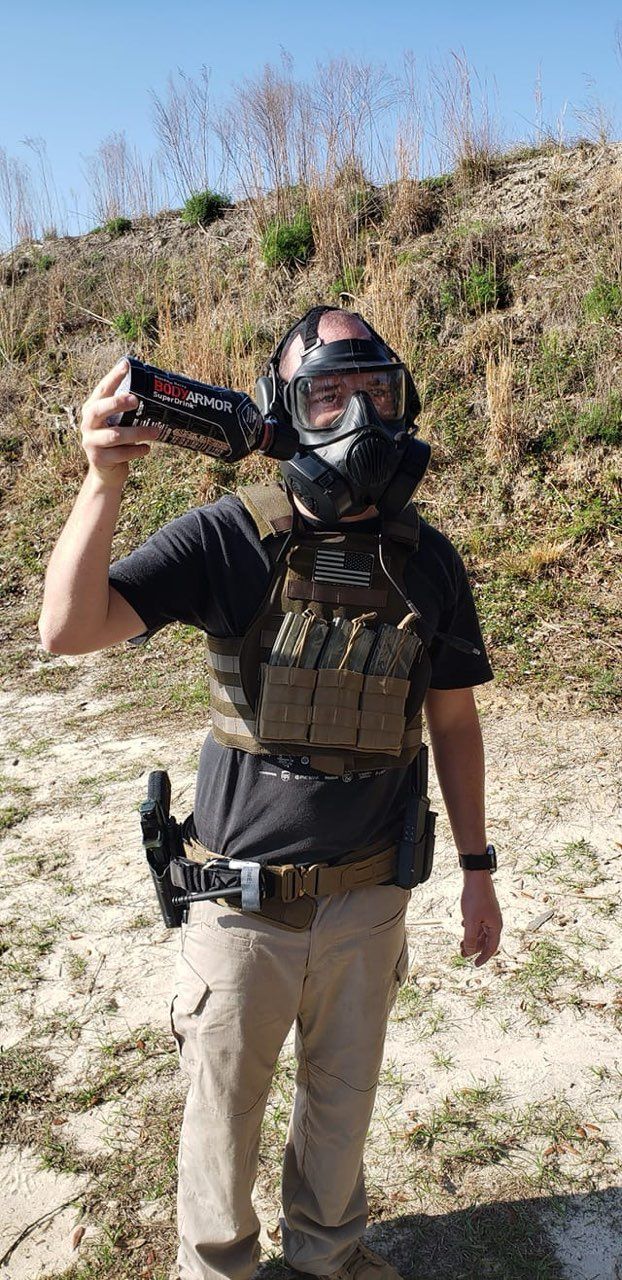 A man in tactical gear, including a gas mask, holding an item. Standing outdoors, sunny day, background.
