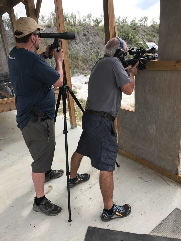 Two men shooting rifles at an outdoor range. One uses a spotting scope.