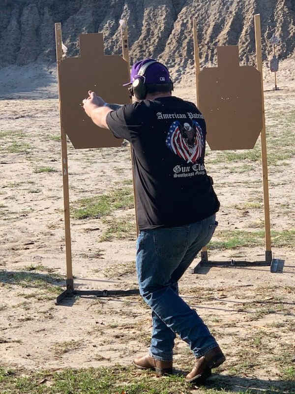 Man shooting at targets at an outdoor range; wearing purple hat, jeans, black t-shirt with American flag design.