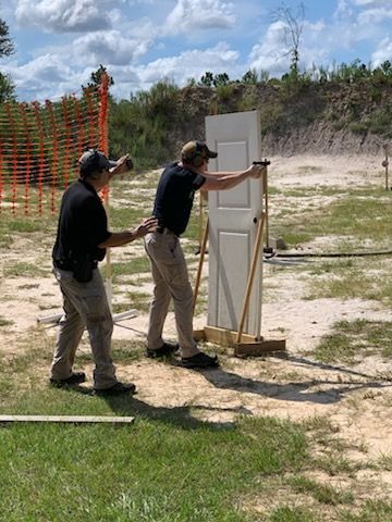 Two men practicing gun shooting at an outdoor range. One is aiming at a door, instructor nearby.