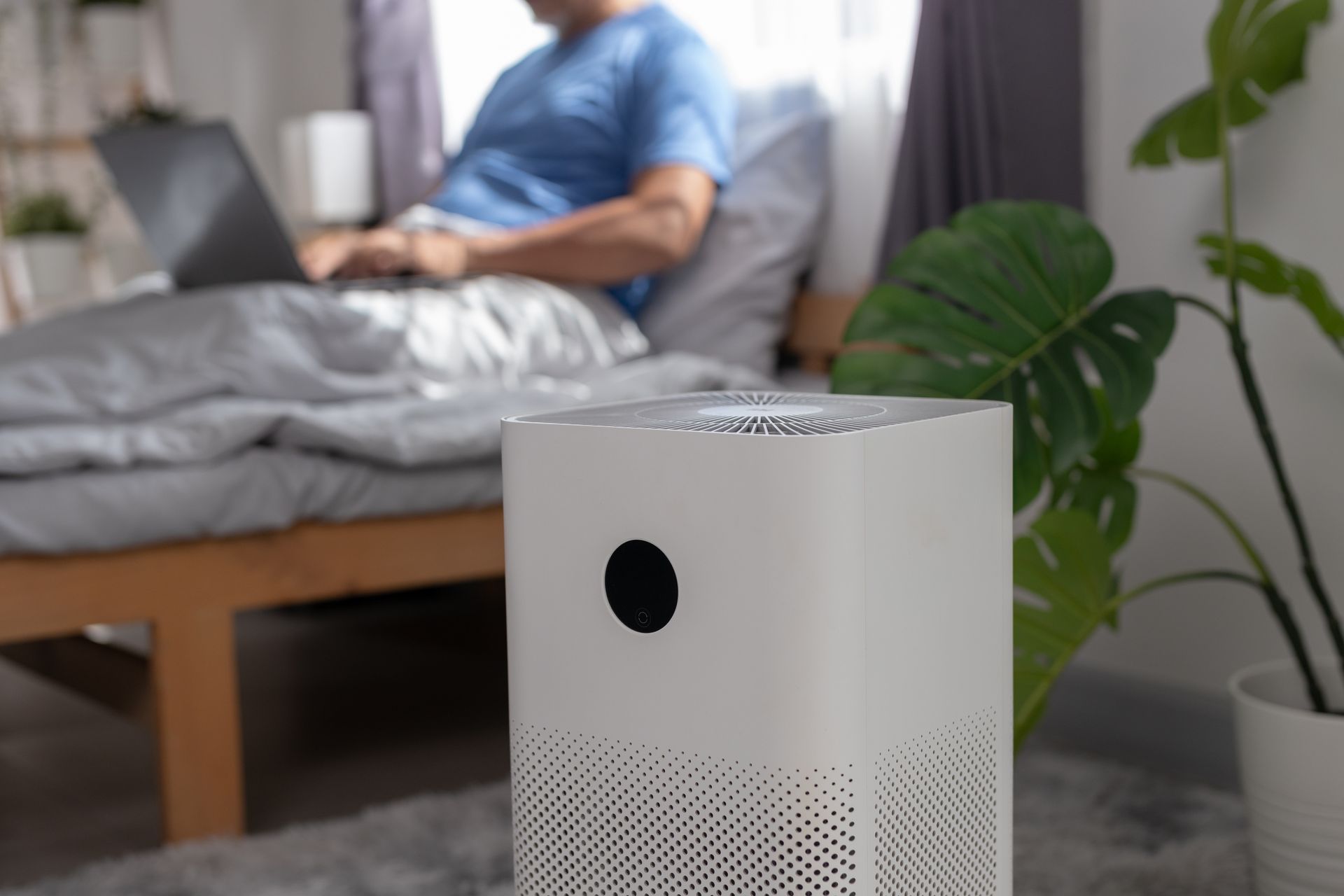 White air purifier in a bedroom with a person using a laptop on the bed in the background White air purifier in a bedroom with a person using a laptop on the bed in the background