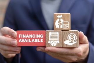 Hands holding wooden blocks and a red block reading “FINANCING AVAILABLE” against a blurred suit background Hands holding wooden blocks and a red block reading “FINANCING AVAILABLE” against a blurred suit background
