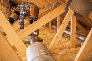 Worker crouching in an attic, inspecting ductwork and wooden beams above insulation Worker crouching in an attic, inspecting ductwork and wooden beams above insulation