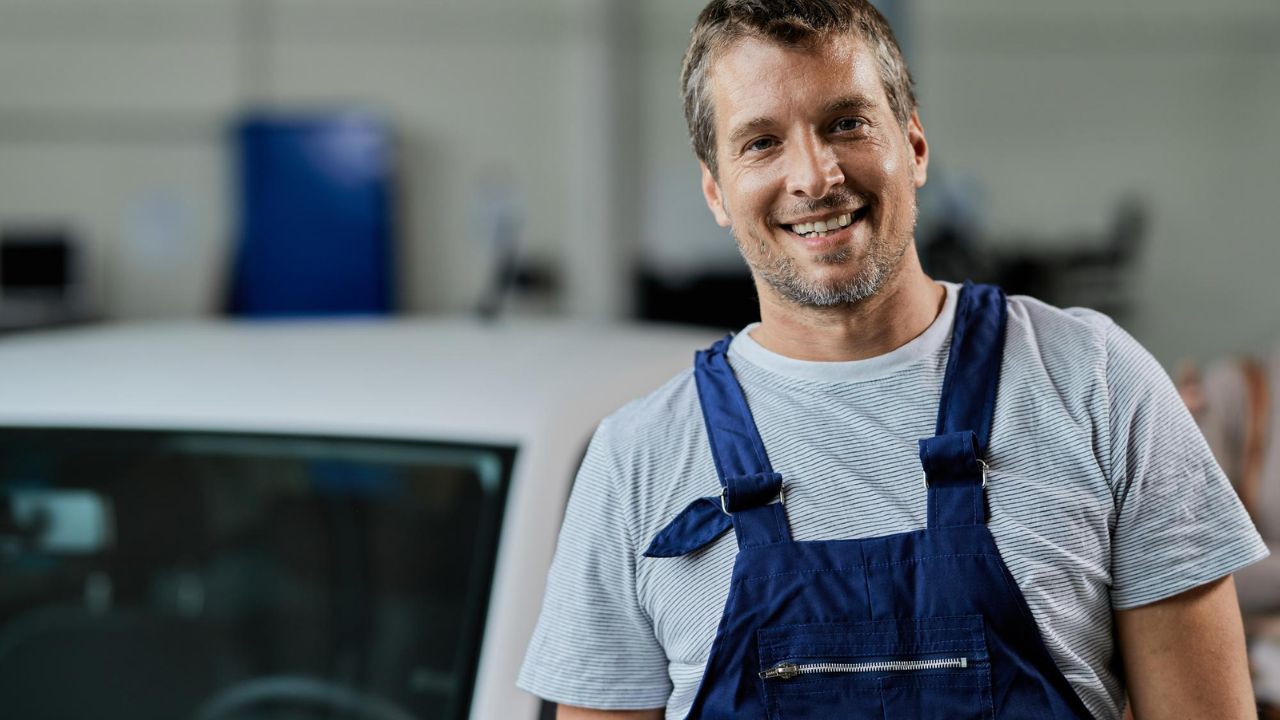 Professional auto mechanic smiling inside an automotive repair shop, ready to provide vehicle maintenance and repair services.