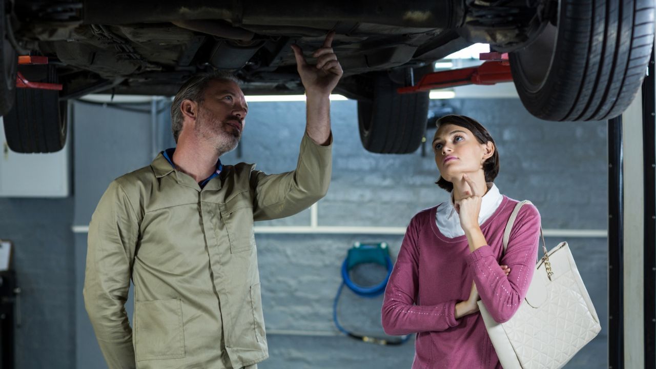 Auto mechanic explaining undercarriage inspection results to a customer while checking a vehicle on a service lift.
