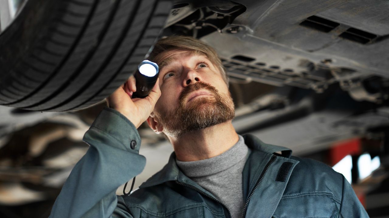 Certified auto mechanic inspecting a car’s undercarriage with a flashlight during a detailed vehicle safety inspection.