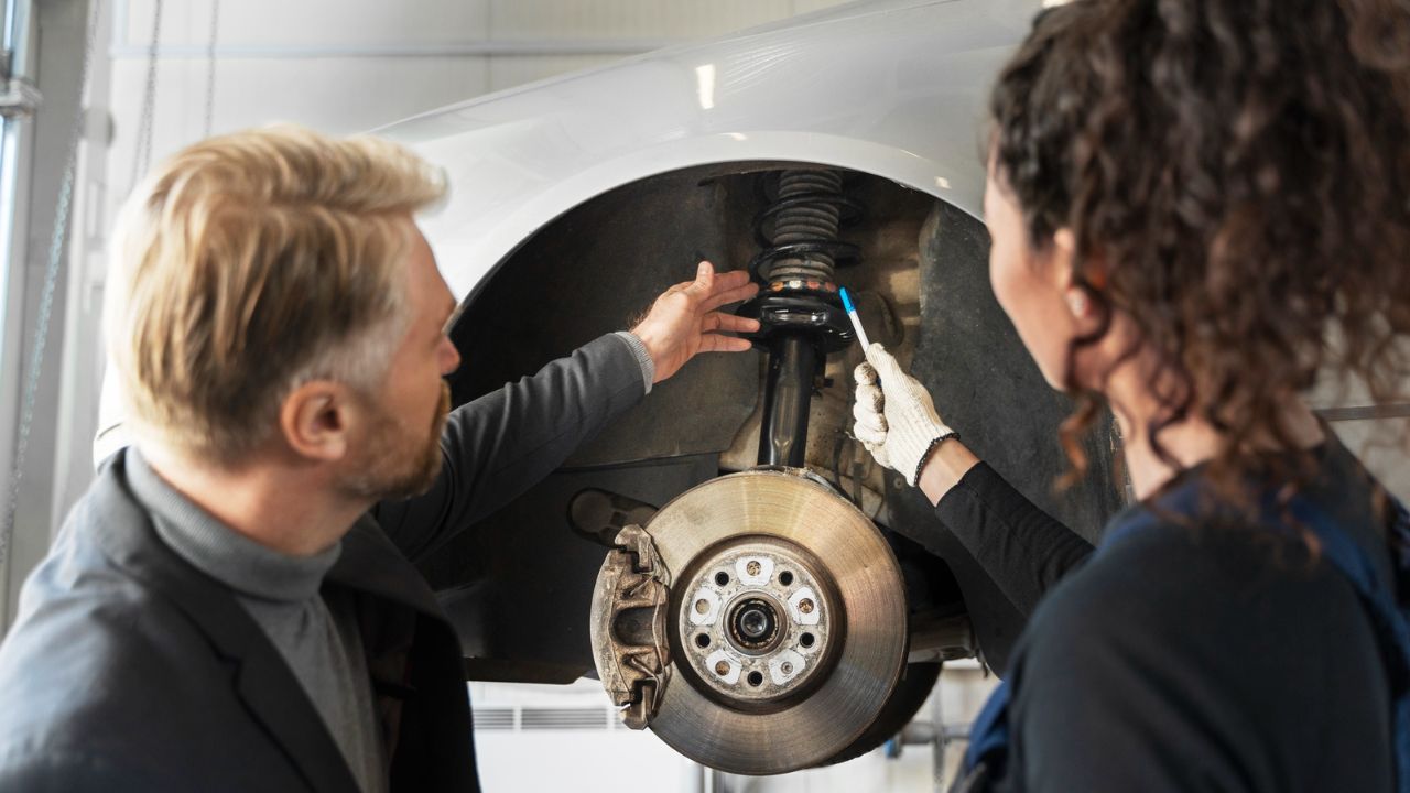 Two people examining a car’s suspension strut above an exposed brake rotor and caliper.