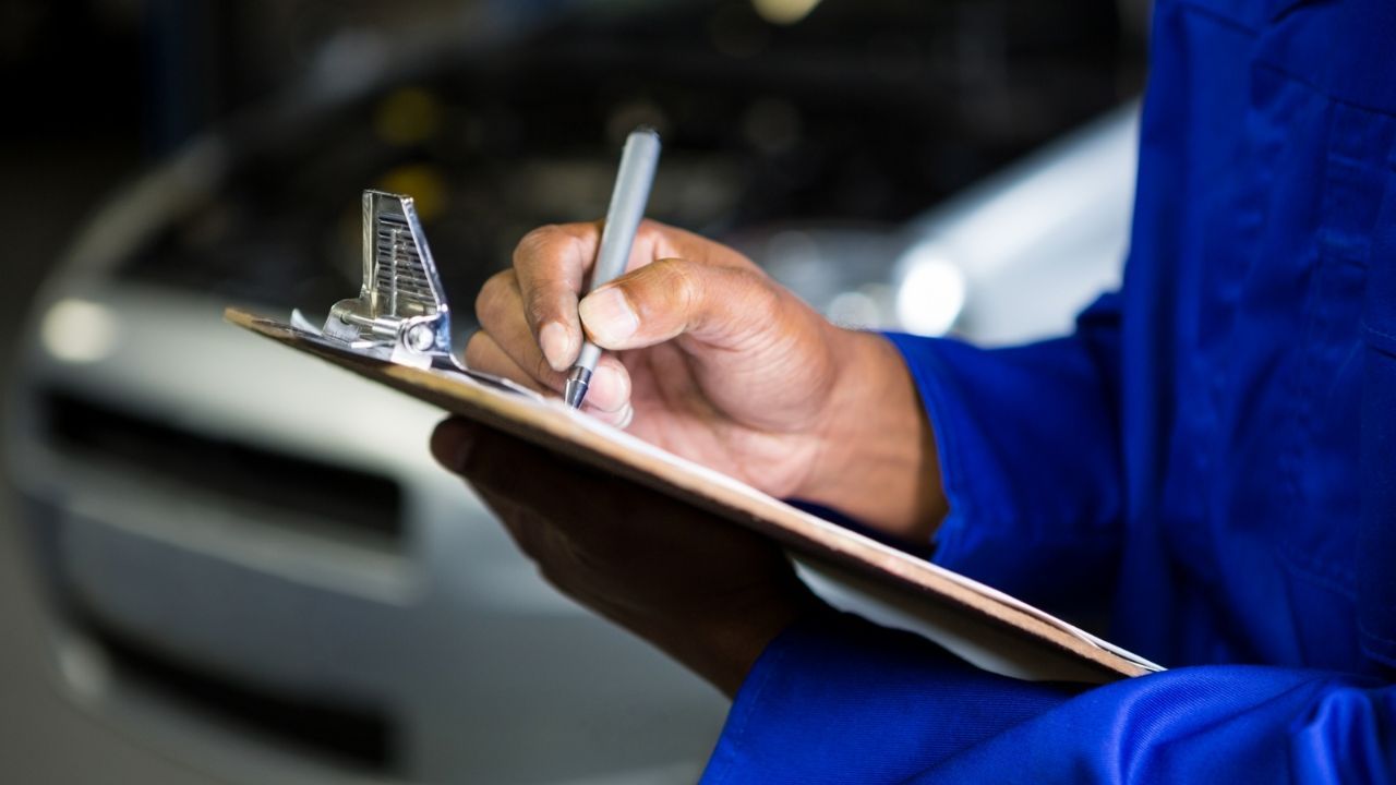 Close-up of a technician in blue workwear writing diagnostic notes on a wooden clipboard in front of an open car hood.