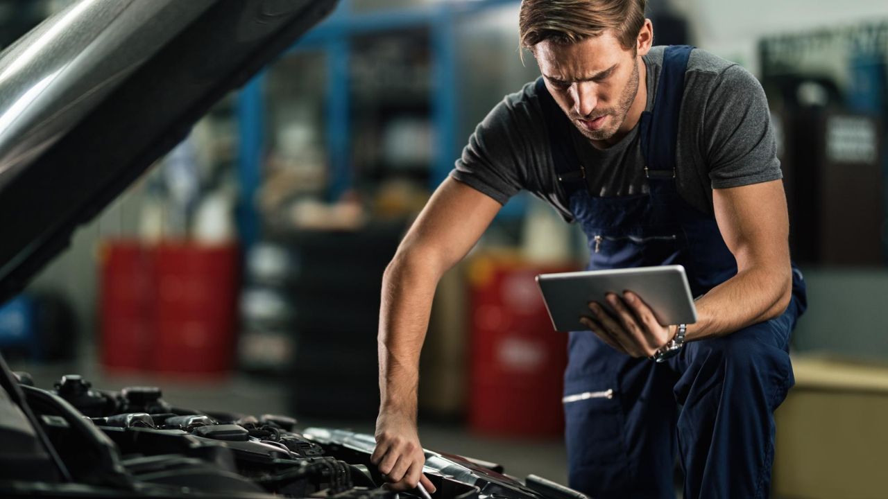 Male mechanic holding a digital tablet while performing an engine inspection on a vehicle in a repair shop.