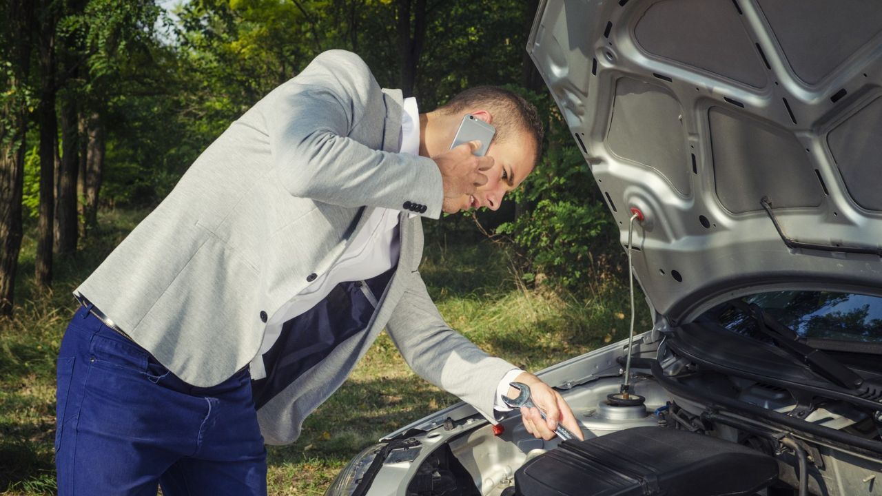 Driver checking the engine under an open hood while talking on a smartphone roadside.