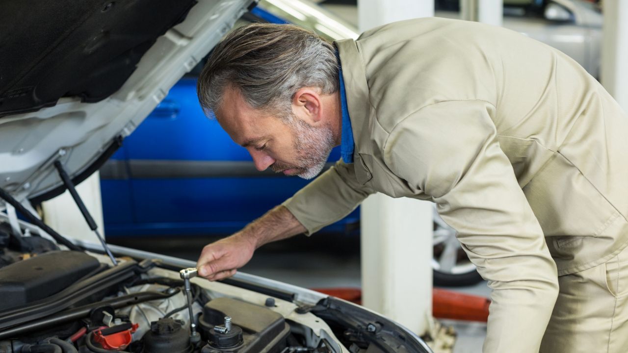Driver checking the engine under an open hood while talking on a smartphone roadside.
