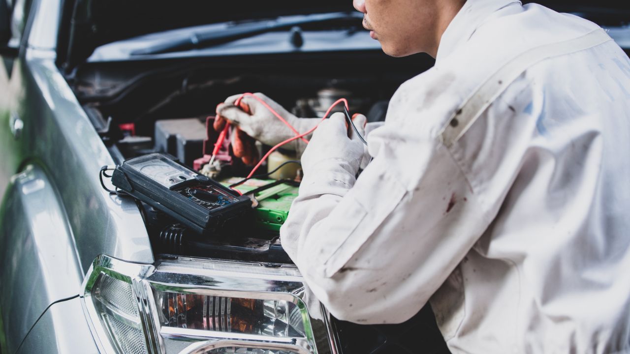 Auto repair technician testing a car battery with a multimeter to diagnose battery and charging system performance.