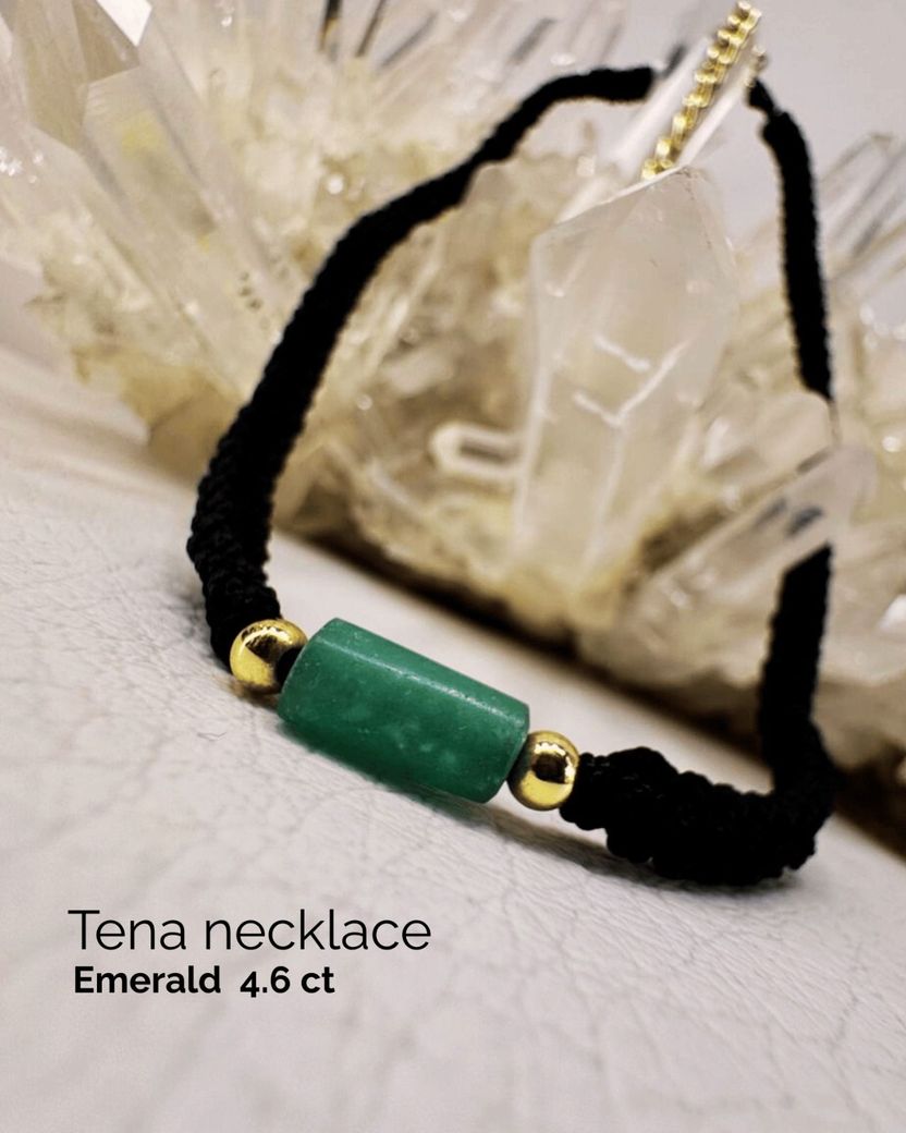 Collar de cordón negro con colgante de esmeralda verde y detalles dorados, exhibido sobre una superficie clara.