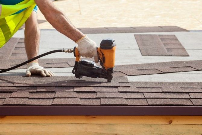 A worker in a yellow safety vest uses a pneumatic nail gun to install brown asphalt roofing shingles on a wooden roof.