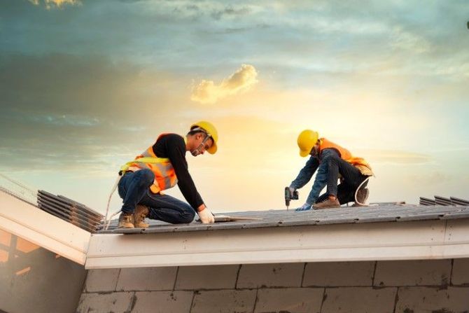 Two construction workers in safety vests and hard hats install shingles on a roof at sunset.