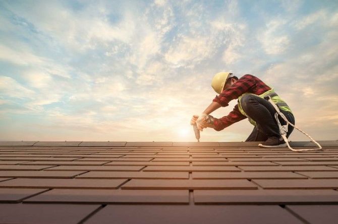 A roofer wearing a hard hat and safety harness uses a power drill on a tiled roof at sunrise.