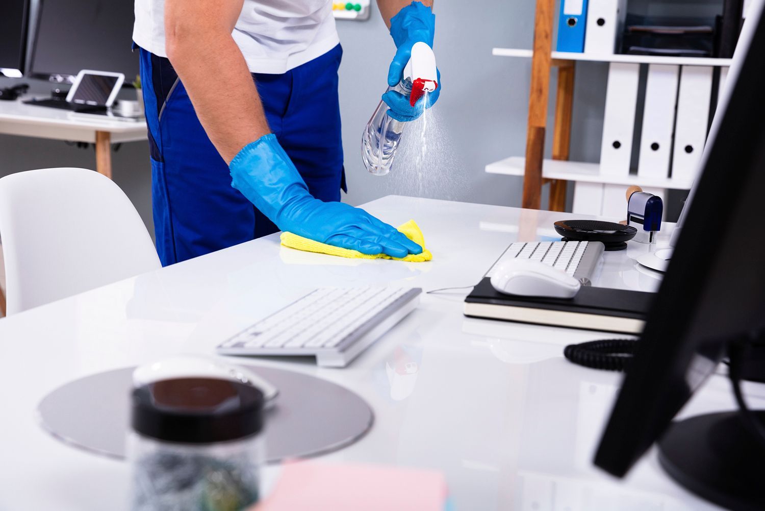Person wearing blue gloves, cleaning a white office desk with spray and a yellow cloth. Person wearing blue gloves, cleaning a white office desk with spray and a yellow cloth.