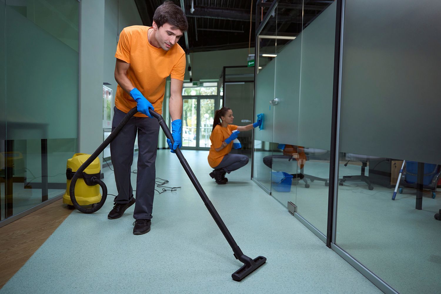 Two people cleaning an office: one vacuuming the floor, the other wiping glass, both wearing gloves and orange shirts.