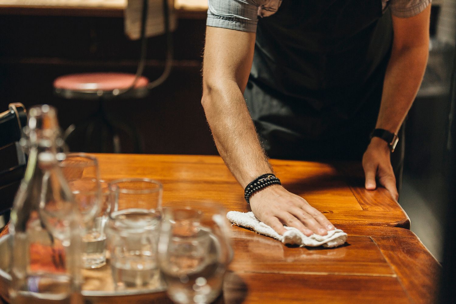 Person wiping a wooden table in a bar; glasses and a stool are visible. Person wiping a wooden table in a bar; glasses and a stool are visible.