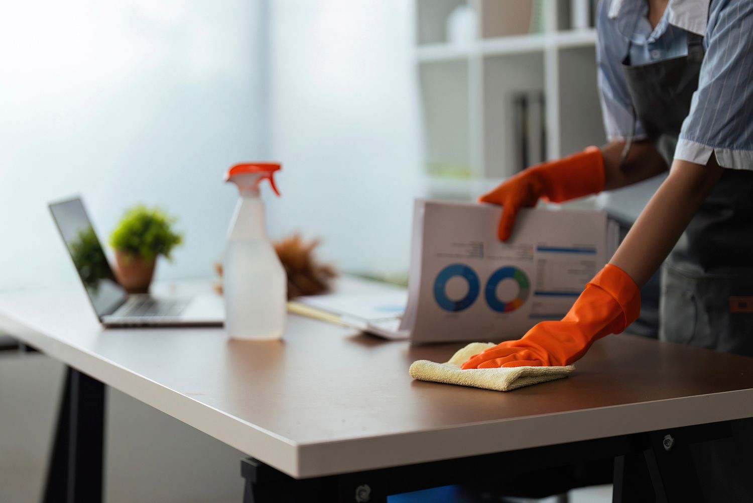 Person in orange gloves cleaning a desk in an office. A laptop, spray bottle, and papers are on the desk. Person in orange gloves cleaning a desk in an office. A laptop, spray bottle, and papers are on the desk.