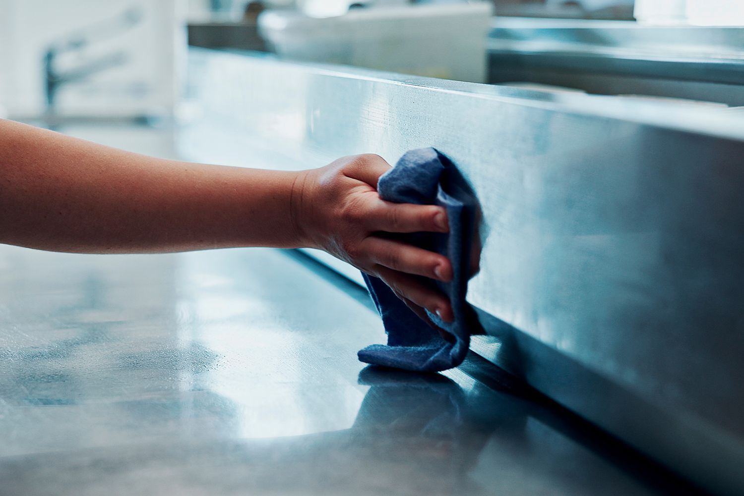 Hand wiping down a stainless steel countertop with a blue cloth. Hand wiping down a stainless steel countertop with a blue cloth.