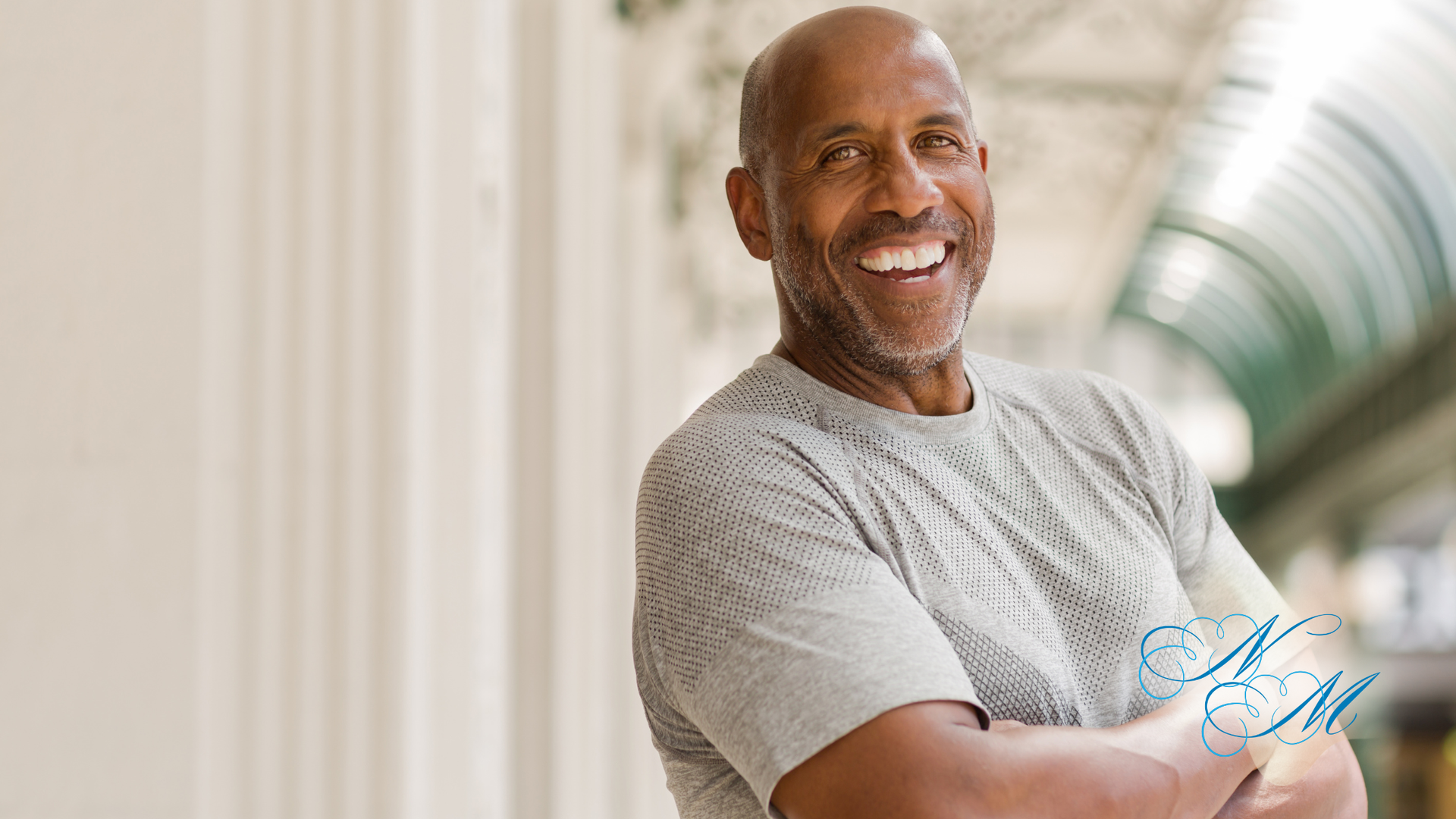 Man smiling, arms crossed. Wearing gray shirt, standing outdoors with white columns and green ceiling in background.