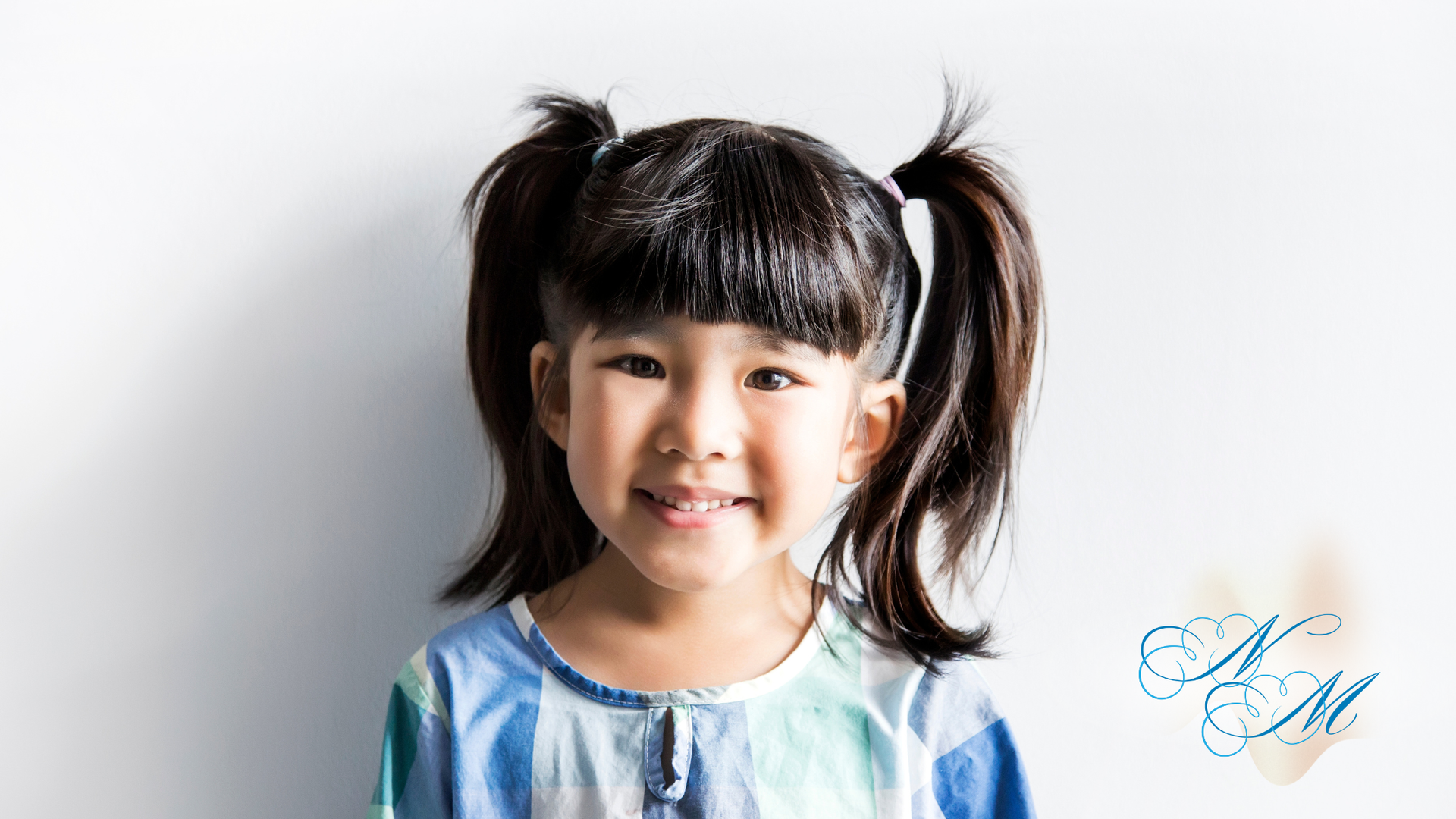 Smiling child with dark hair in pigtails and bangs, wearing a blue and white striped shirt, standing against a white wall.