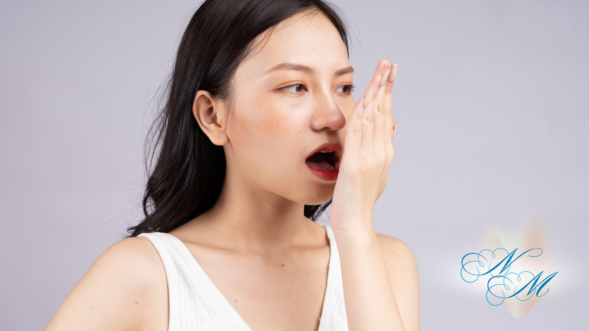 Woman with hand covering mouth, appears to be checking breath. Light background, wearing white top.
