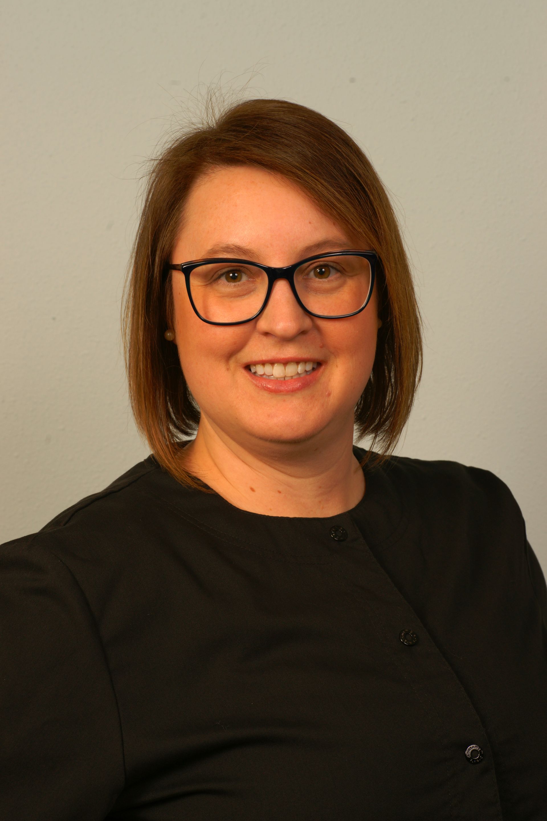 Woman with glasses smiles; wearing a black shirt, posing in front of a gray background.