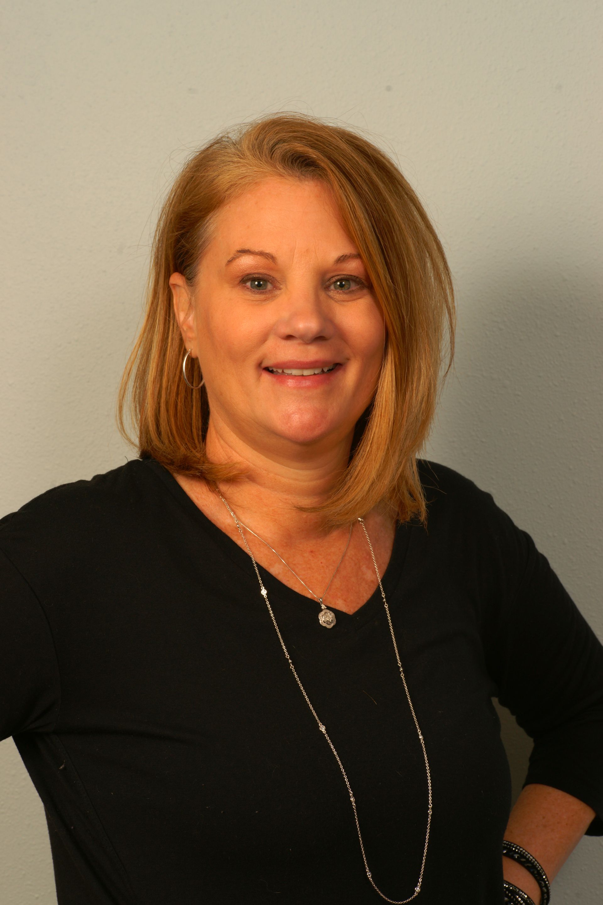 Woman with shoulder-length blonde hair, wearing a black v-neck shirt, and a long silver necklace, smiling.