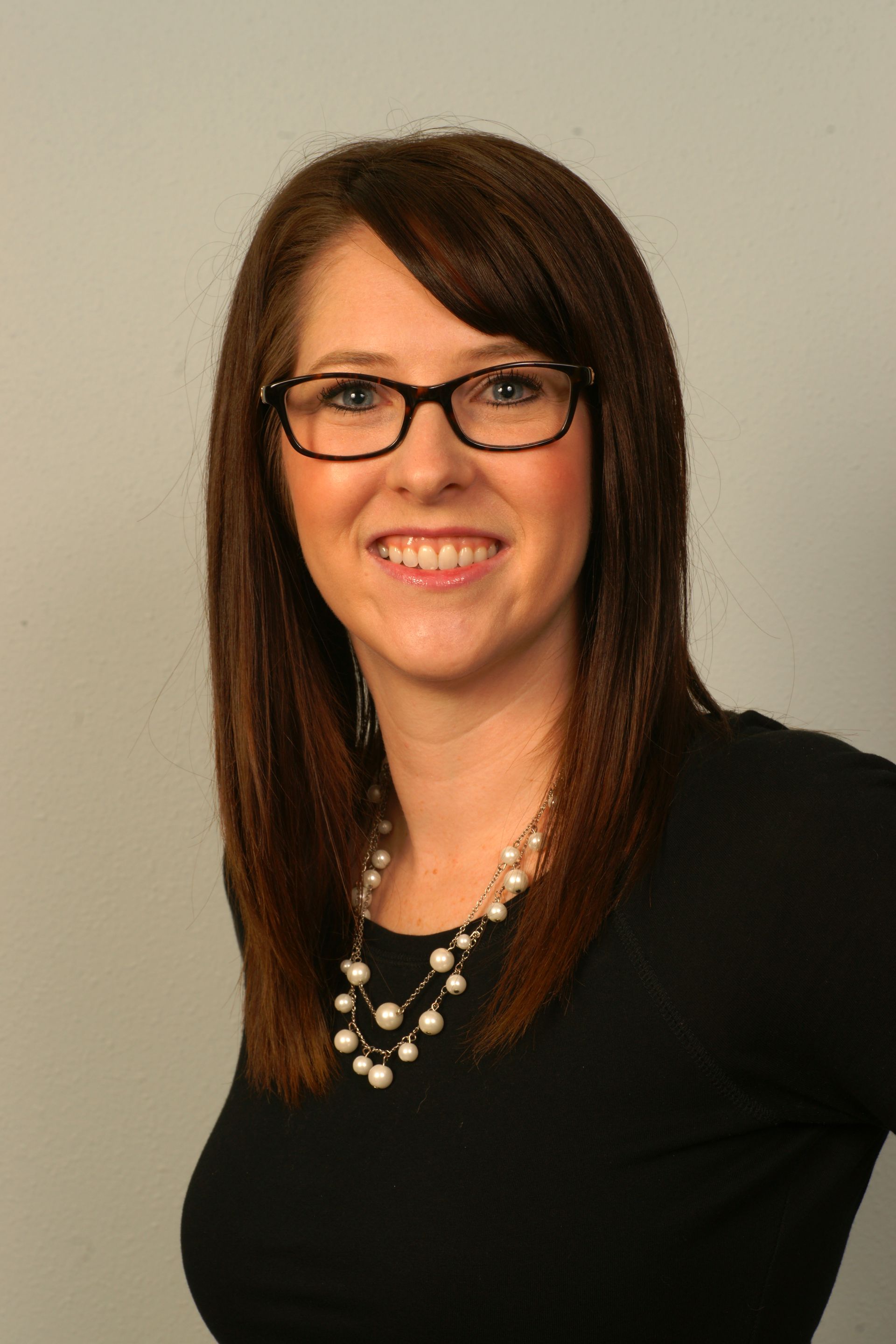 Woman with glasses and pearl necklace smiles, posing against a grey backdrop.