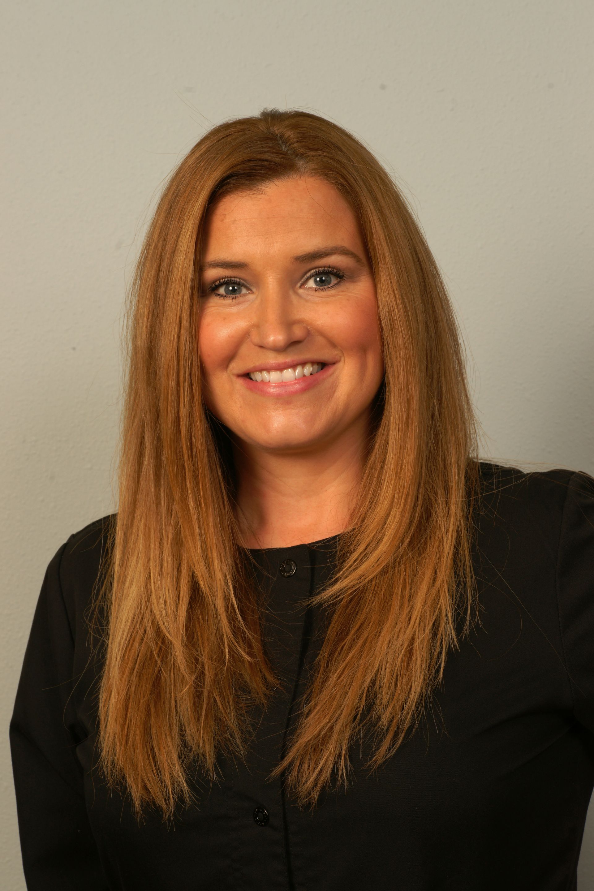 Woman with long auburn hair smiles at the camera, wearing a black shirt against a grey backdrop.