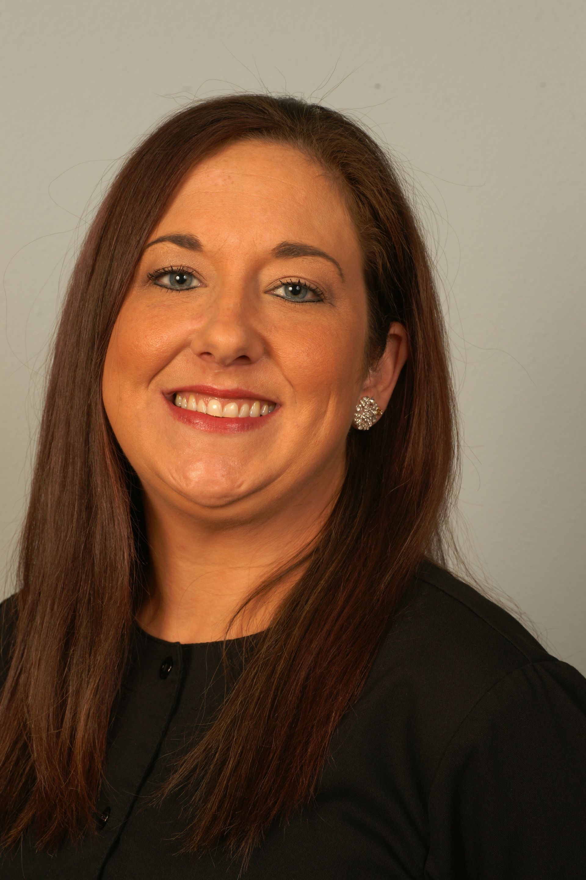 Woman with brown hair, smiling, wearing a black top, against a gray background.