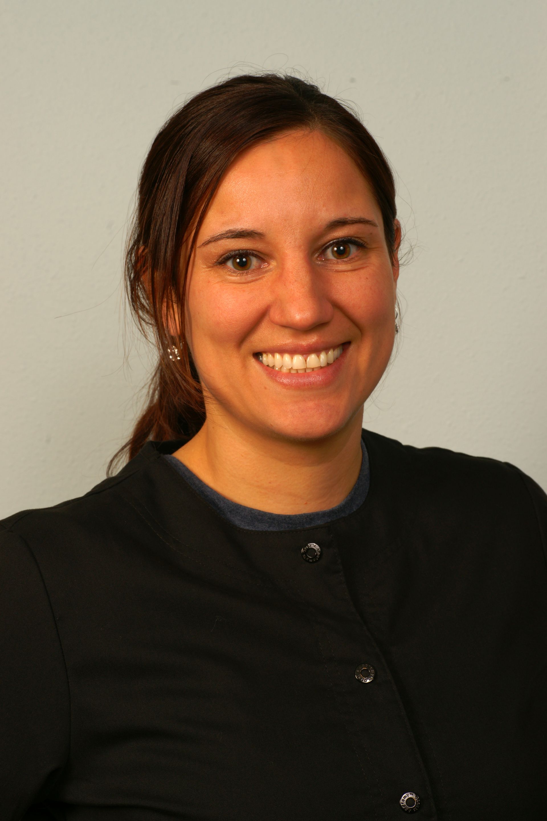 Woman with brown hair smiles, wearing a black cardigan over a blue top, against a grey background.