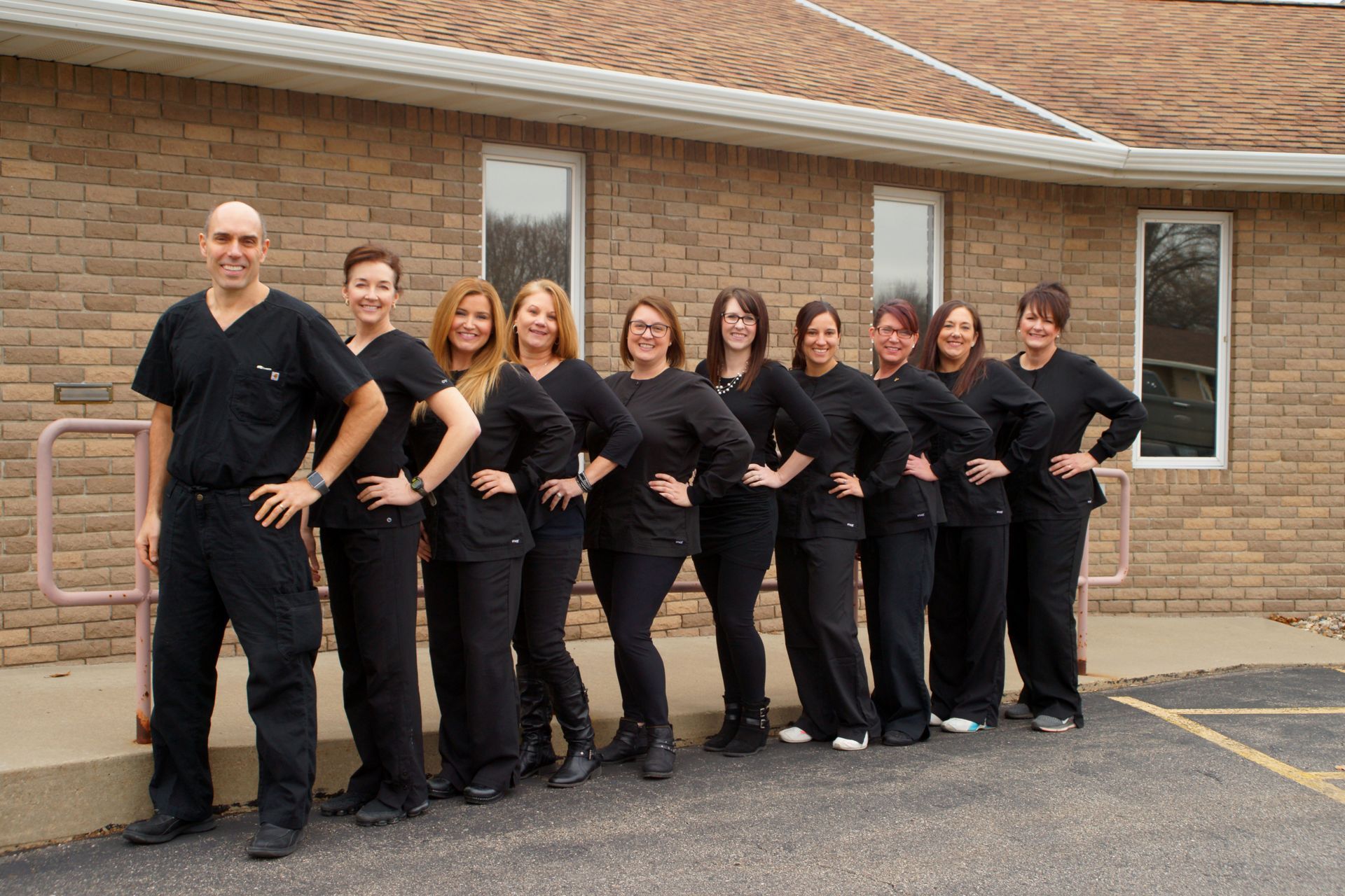 A group of ten people wearing black scrubs standing outside a brick building, posing with hands on hips.