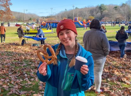 A person in a red beanie and blue fleece holds a pretzel and a cup outdoors.