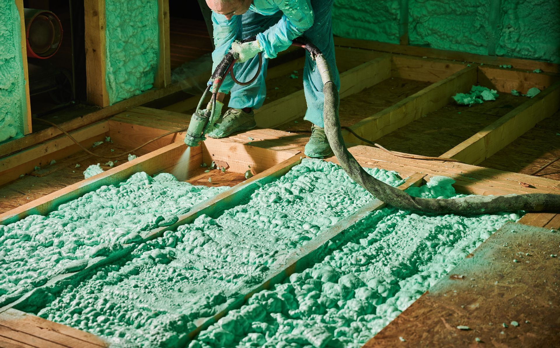 Technician applies spray foam insulation between floor joists inside a building during construction.