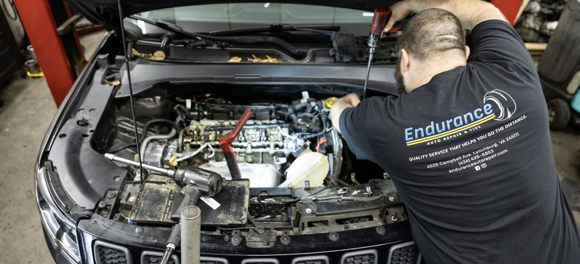 Mechanic working on a car engine in a garage, with the hood open and tools visible. | Endurance Auto Repair and Tire