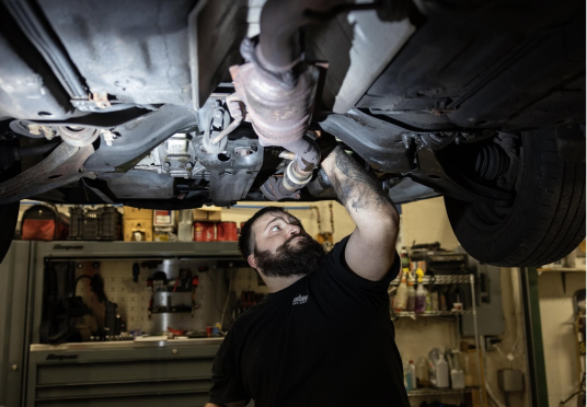 Mechanic inspecting a car’s underside in a garage, reaching up toward the exhaust system. | Endurance Auto Repair and Tire