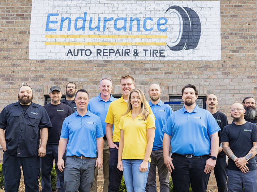Endurance Auto Repair & Tire team posing in front of the shop sign, with two people in bright blue and yellow shirts. | Endurance Auto Repair and Tire
