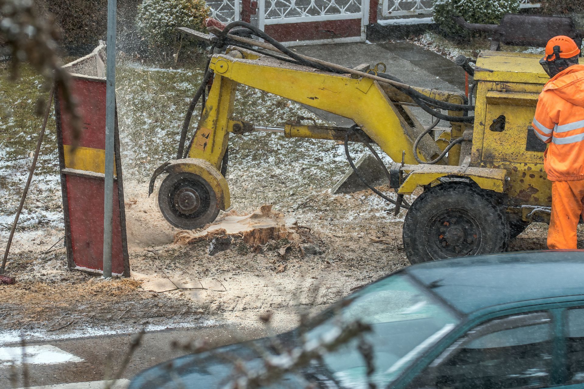 Yellow excavator clearing snow on a street as a worker in orange safety gear stands nearby.